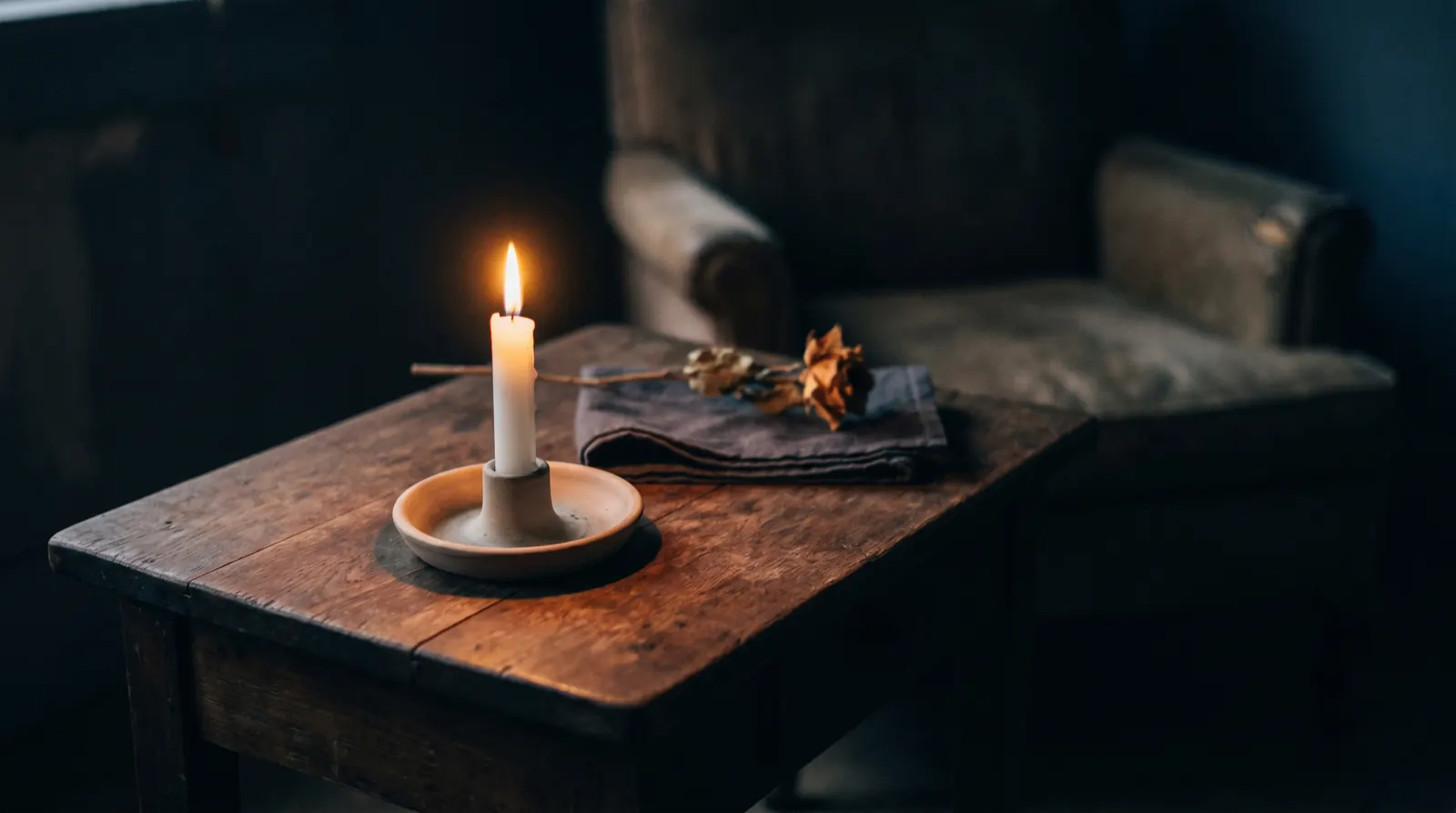 A single lit candle on a wooden table beside a folded cloth and a dried rose